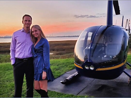 Two people standing outdoors near a helicopter at sunset, smiling, with a grassy area and water in the background.