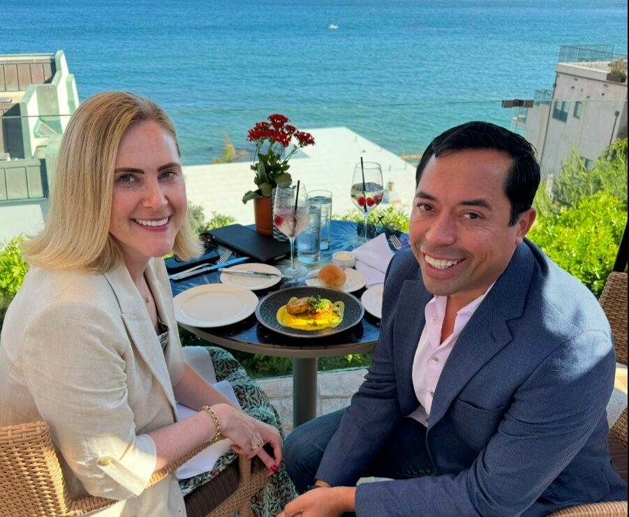 Two people dining outdoors with ocean view, smiling, with a table of food and drinks, greenery, and buildings in background.
