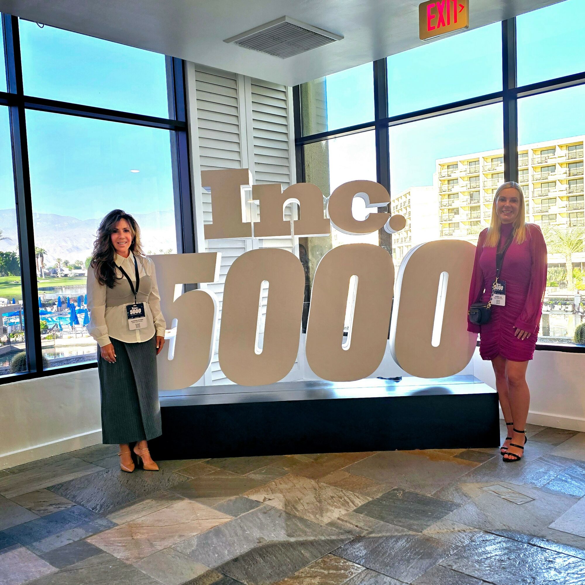 Two women standing beside large 'Inc. 5000' sign inside a building with large windows and city view.