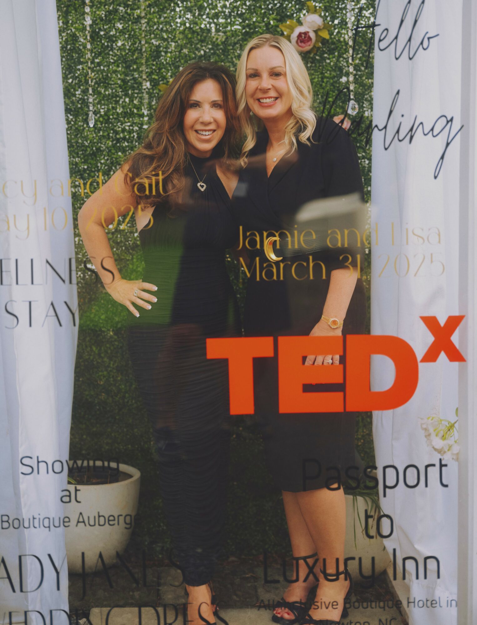 Two women smiling and standing together outdoors with greenery and a white backdrop, wearing black dresses, at a TEDx event.