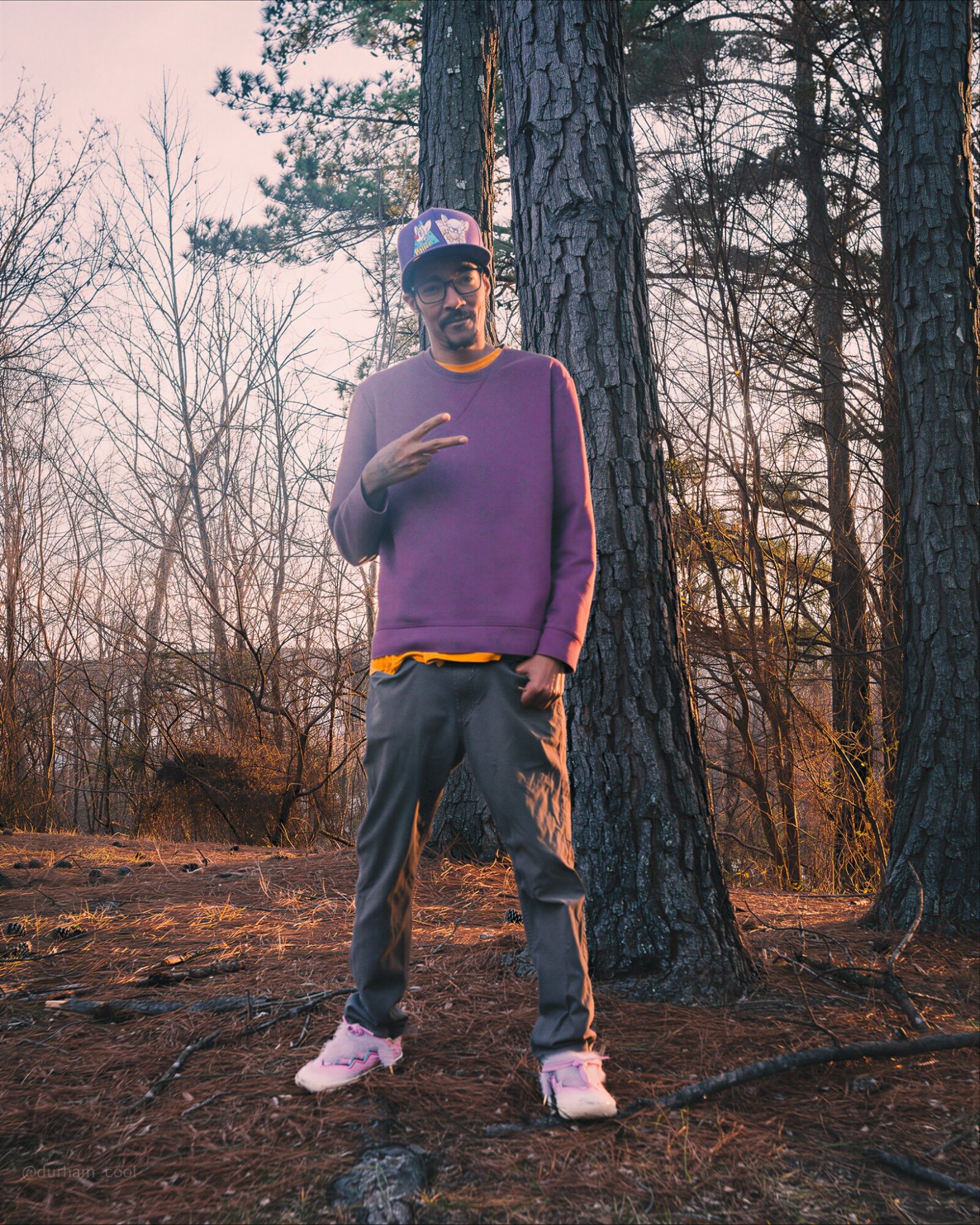 Man standing outdoors in a forested area, wearing a purple sweater, gray pants, and a cap, making a peace sign.