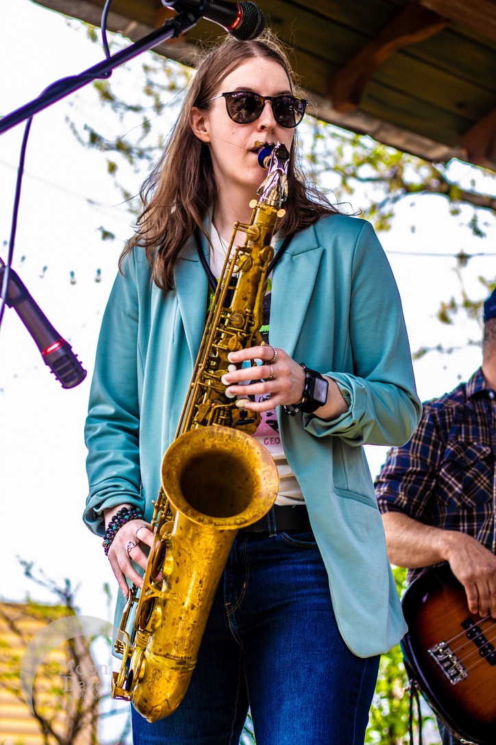 Woman with sunglasses holding a saxophone outdoors, wearing a teal jacket and dark jeans.