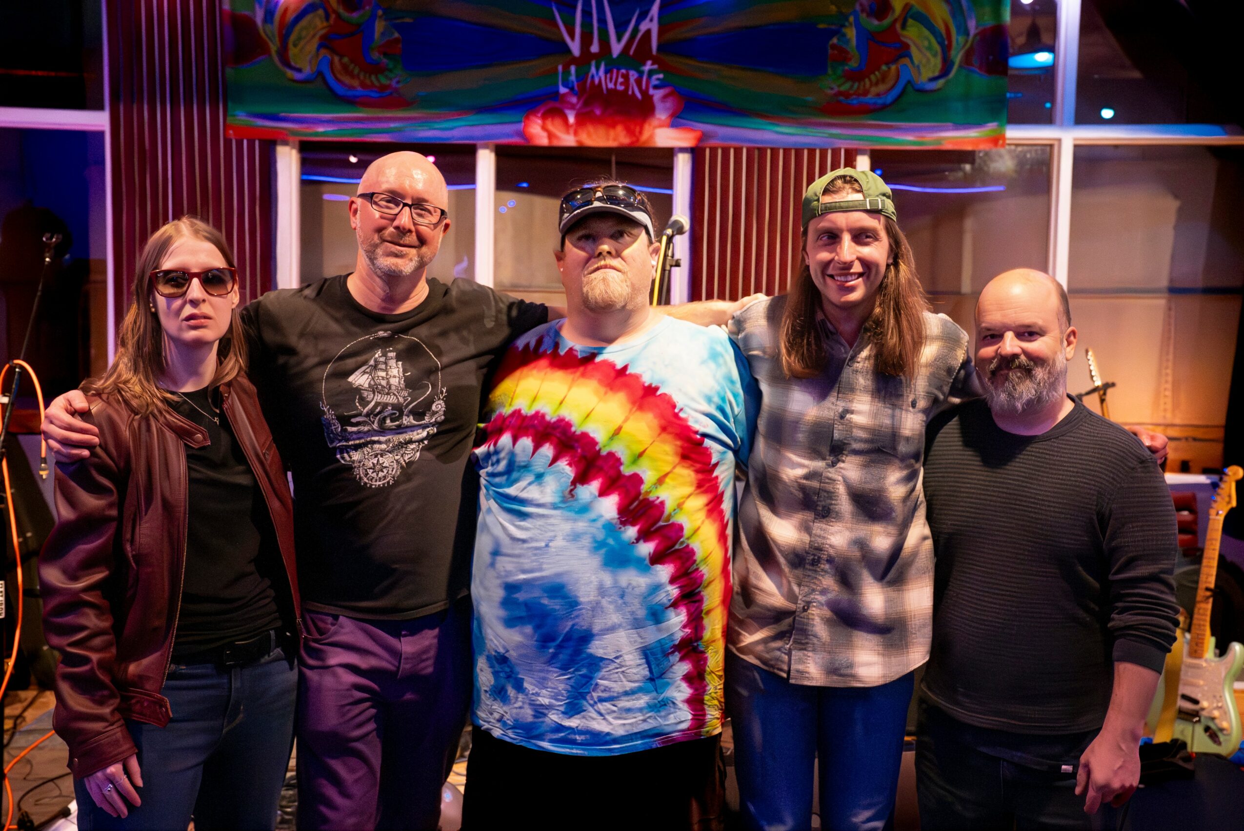 Five people stand together in a colorful room, smiling, with musical instruments and vibrant artwork in the background.