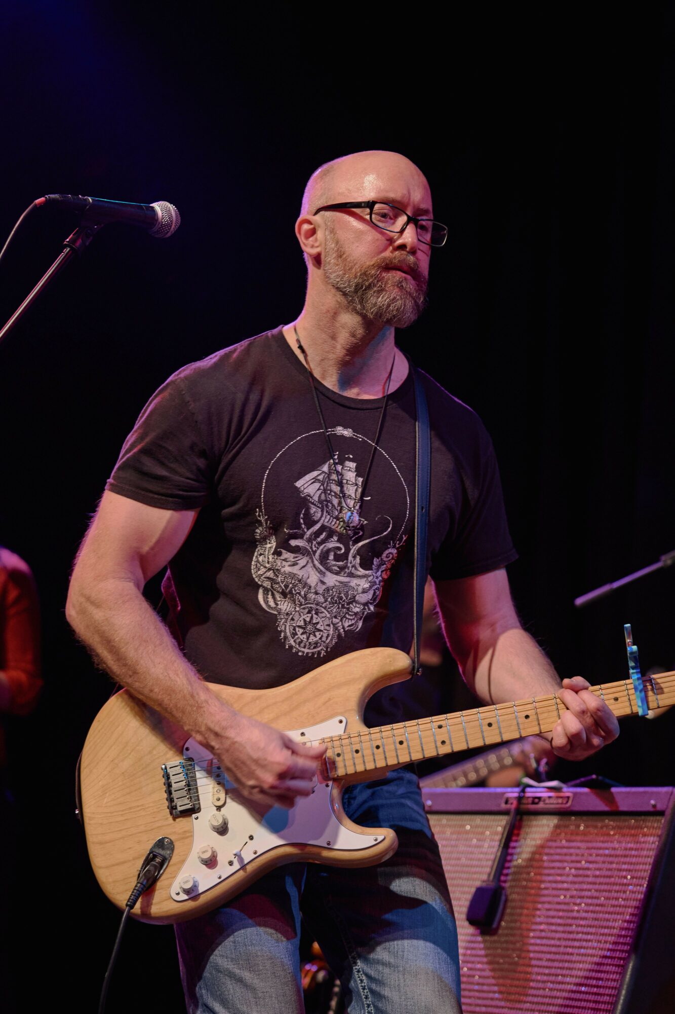 Bald man with glasses playing an electric guitar on stage, wearing a black t-shirt with a design, against a dark background.