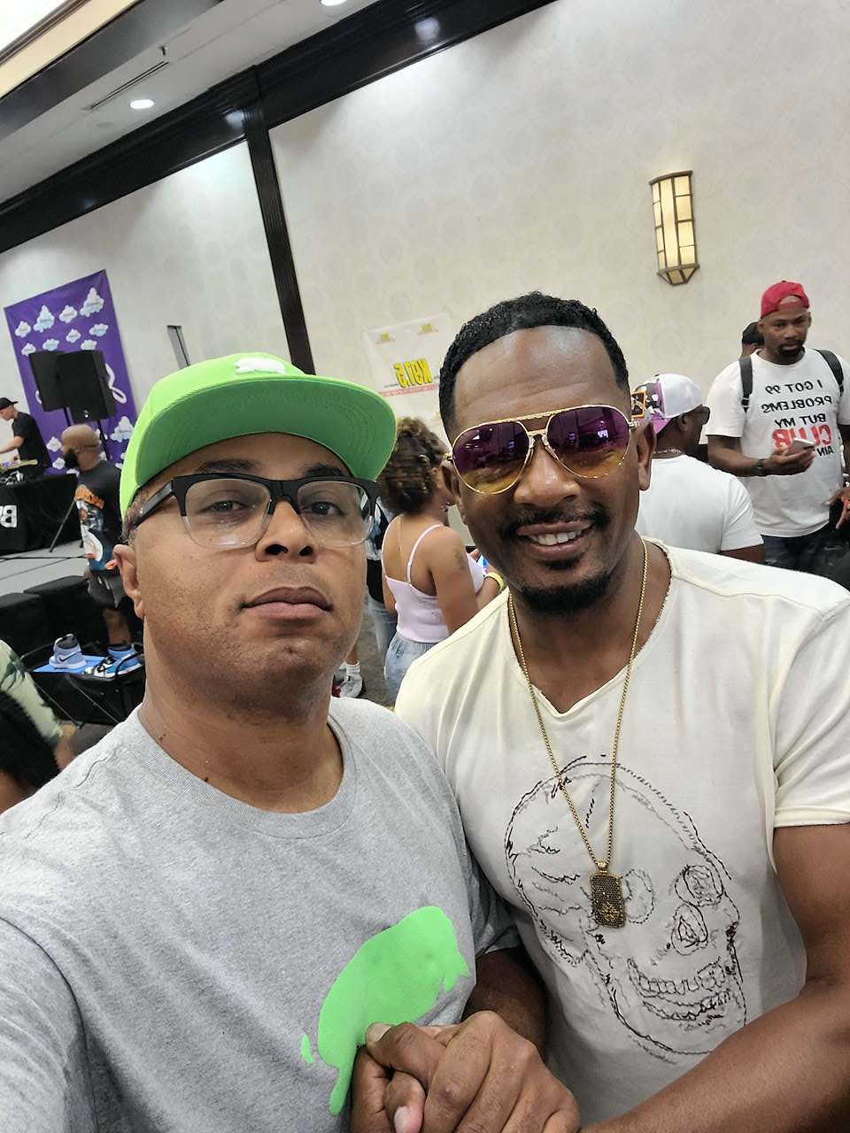 Two men smiling, one wearing sunglasses and a white t-shirt, the other wearing glasses and a green cap, in a crowded indoor setting.