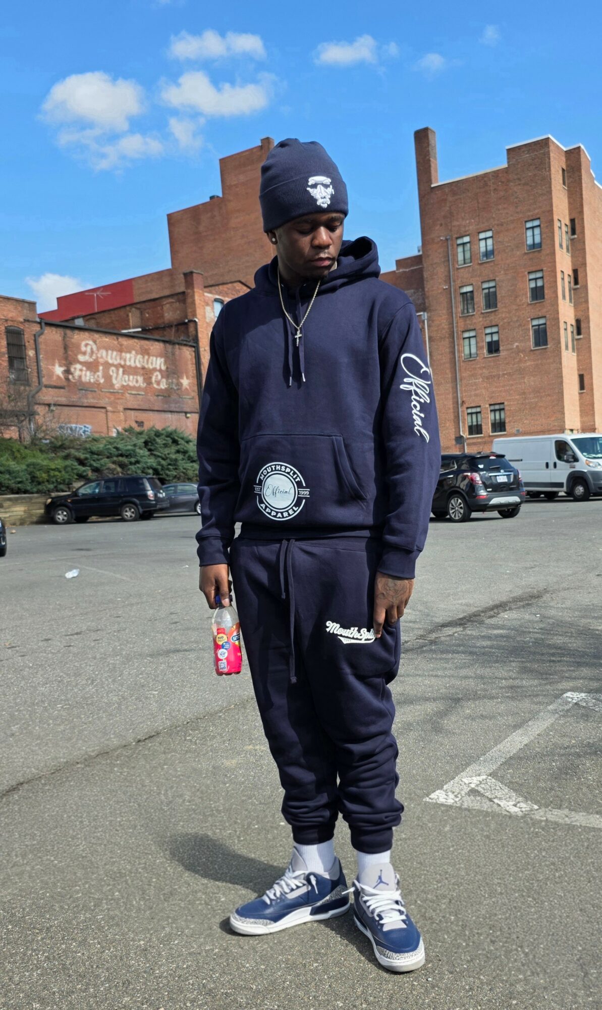 Young man in navy hoodie, sweatpants, and sneakers standing outdoors in a parking lot with brick buildings and cars behind him.