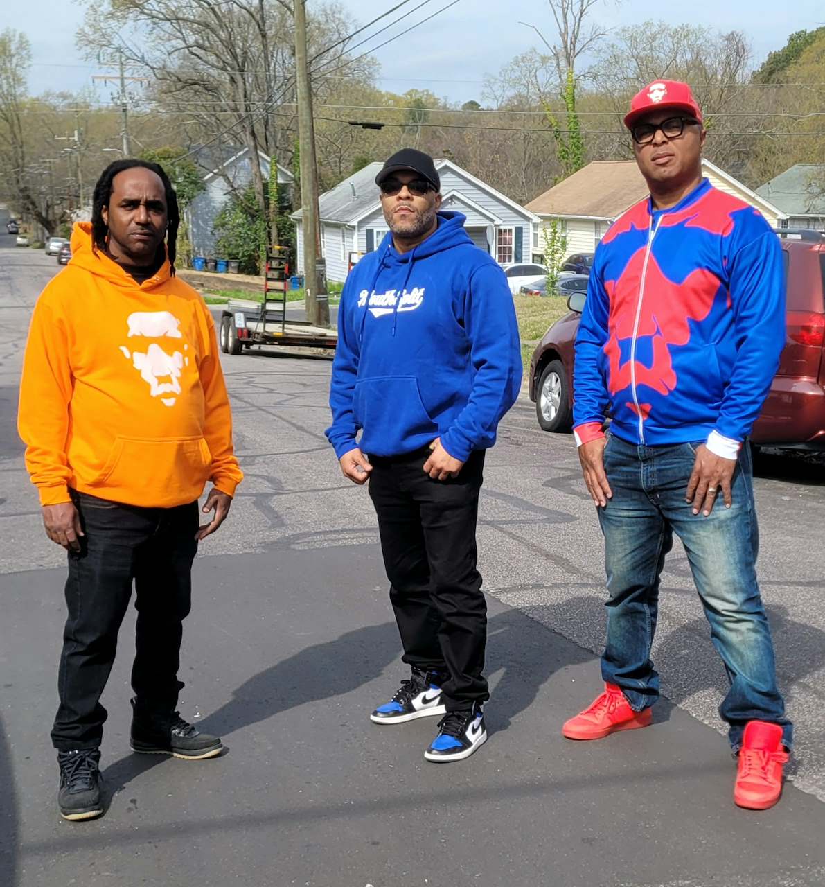 Three men standing outdoors on a street, wearing colorful hoodies and jeans, with houses and trees in the background.