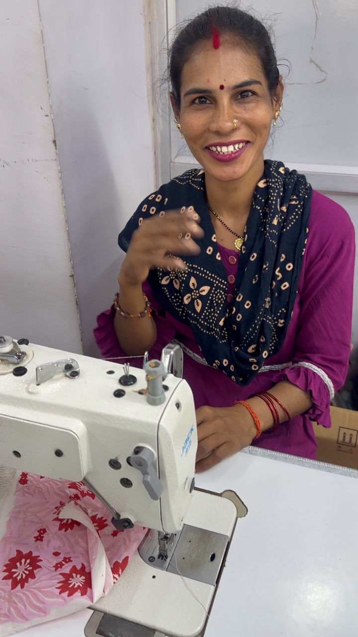 Smiling woman in traditional attire sitting at a table with a sewing machine, fabric, and tools.