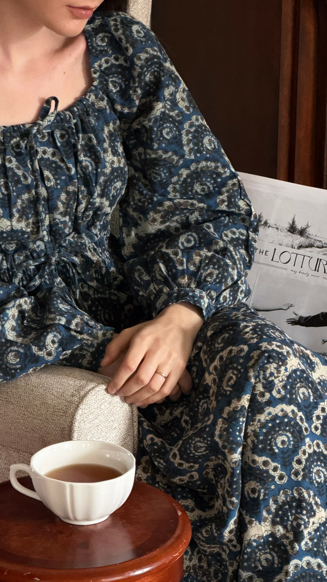 Person sitting with a cup of tea on a wooden table, wearing a patterned blue dress, in a cozy indoor setting.