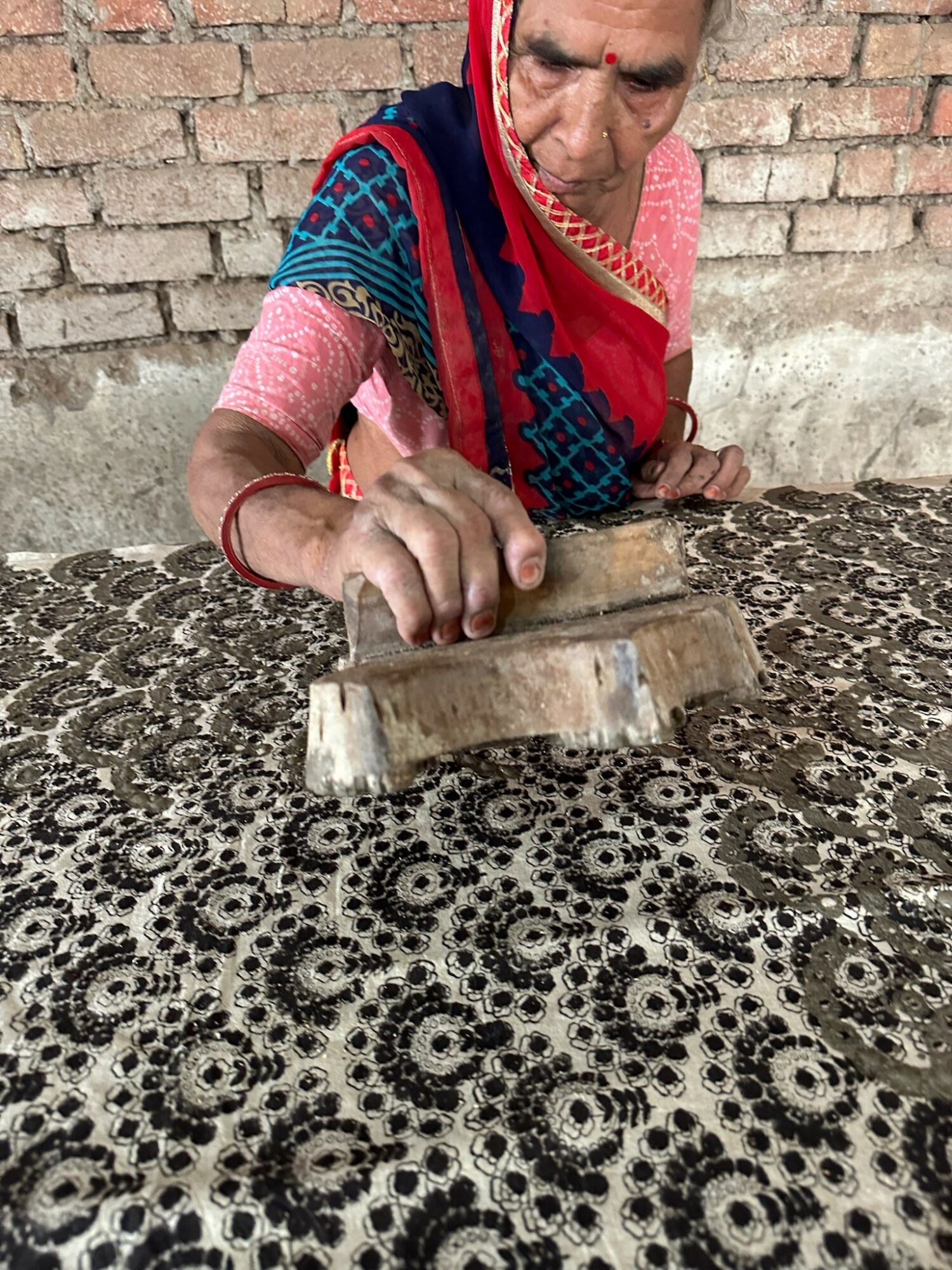 Elderly woman in traditional clothing presses a stamp onto a patterned surface outdoors, with a brick wall background.