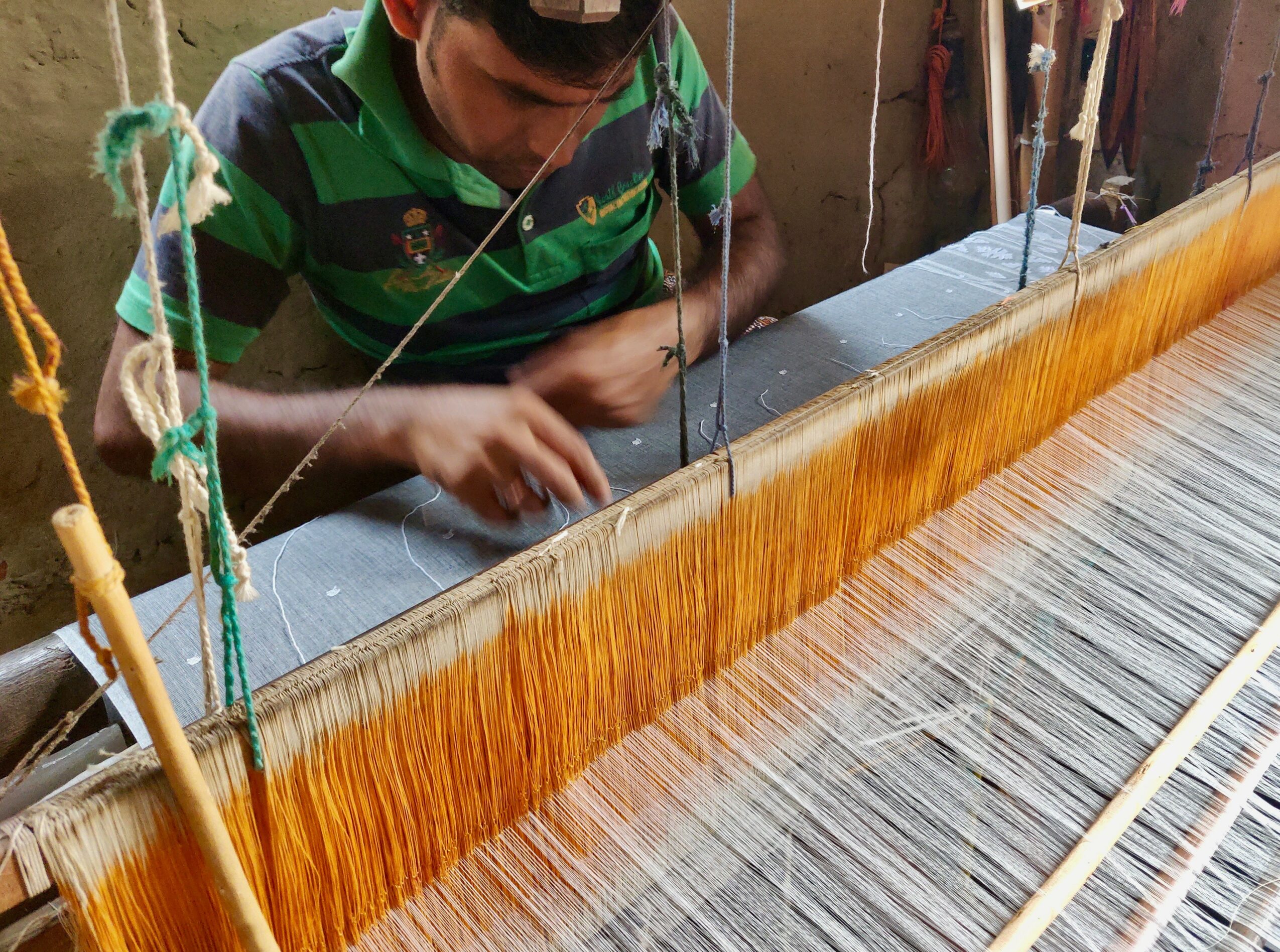 Person working on a traditional loom with orange and gray threads, weaving fabric in a workshop.