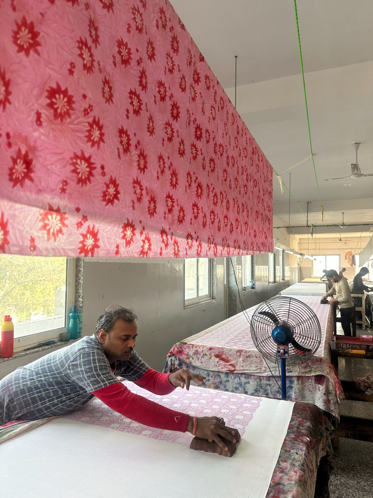 Man working on fabric in a room with tables, fans, and windows, with a pink floral fabric hanging overhead.