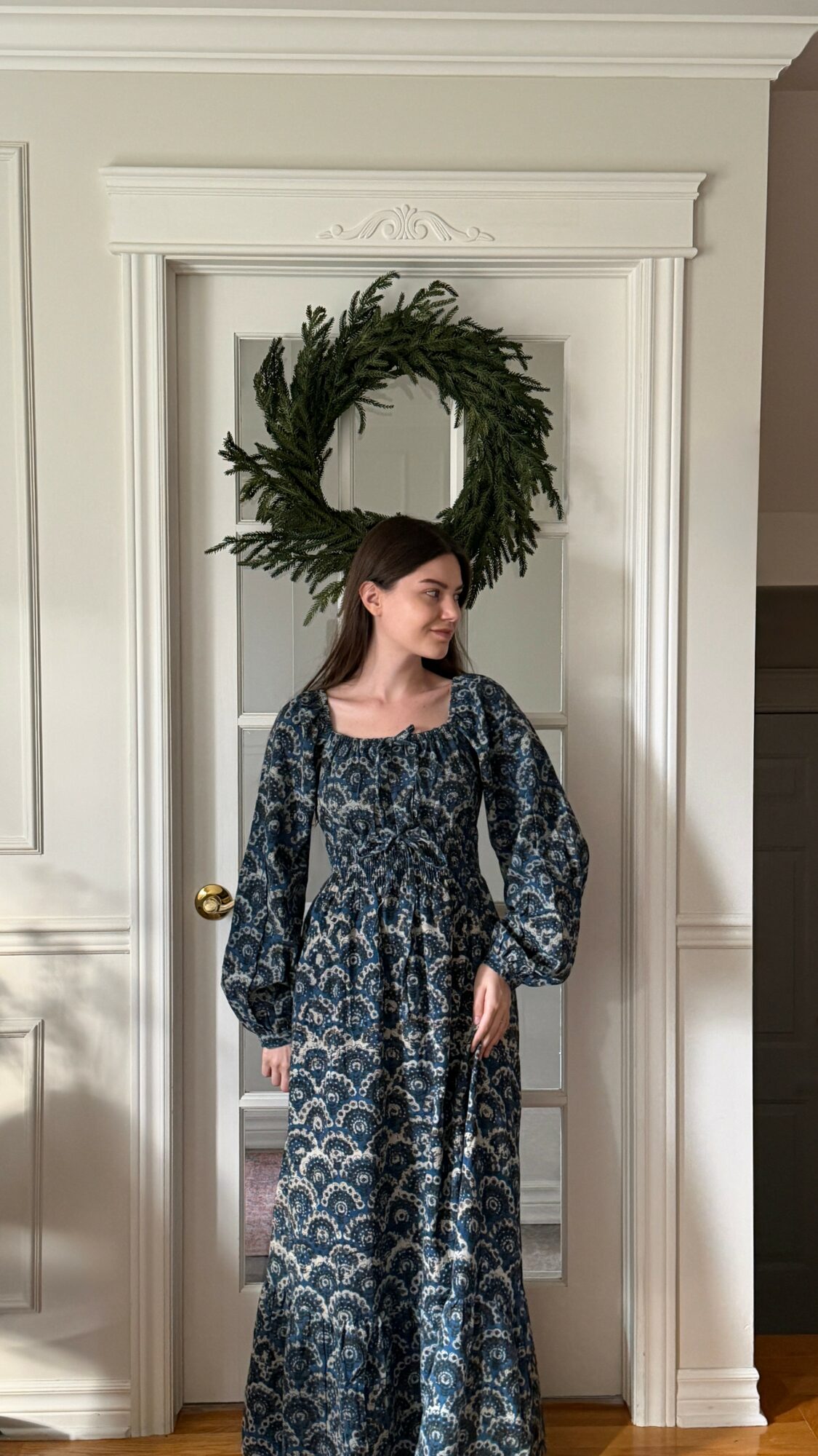 Woman in patterned dress standing in doorway with a wreath above her head, white trim and walls, gold doorknob