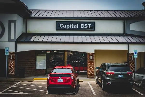 Storefront with a sign reading 'Capital BST', two parked cars in front, and a parking lot with designated spaces.