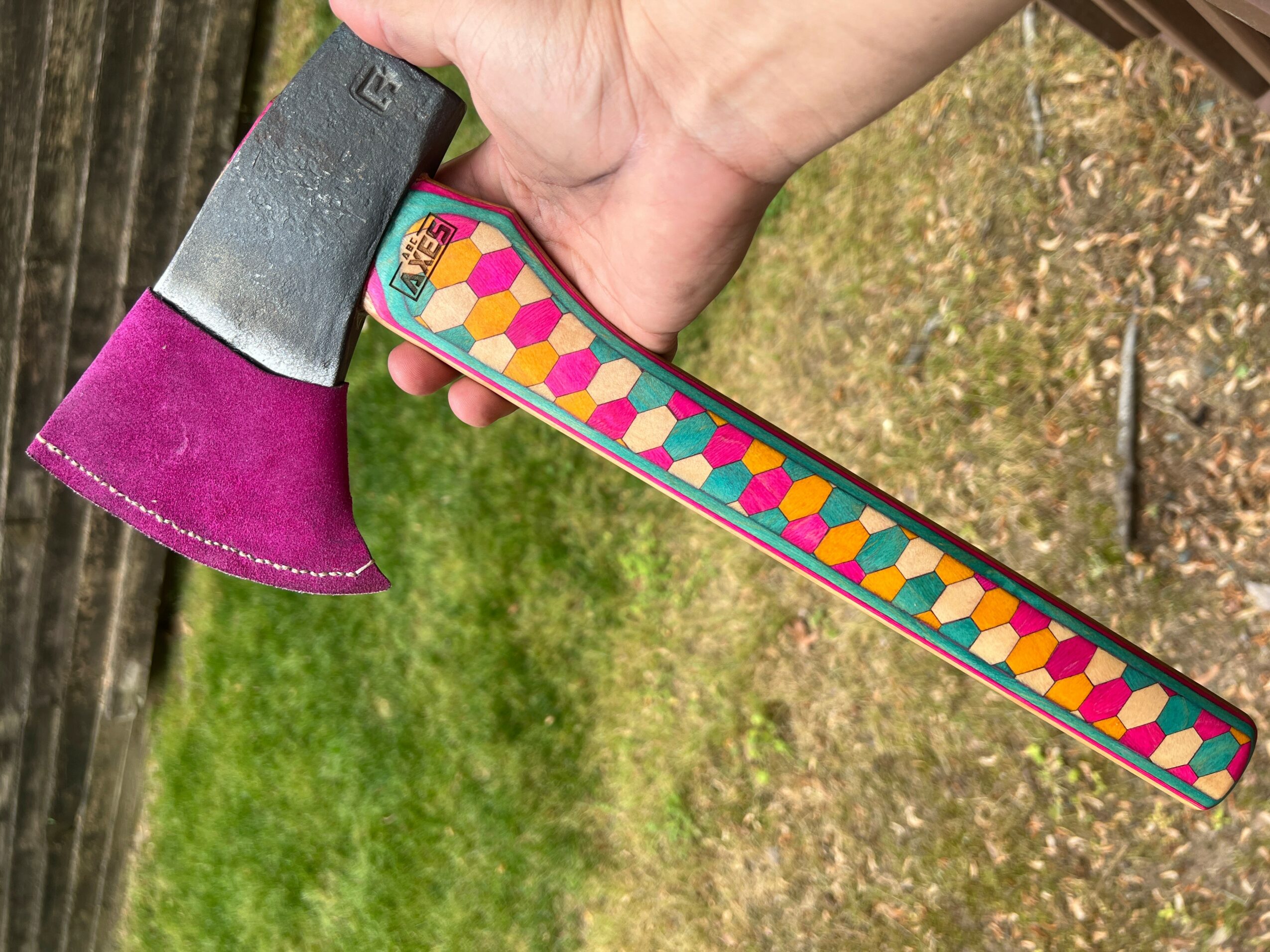 Hand holding an axe with a colorful patterned handle, outdoors on grass and trees.