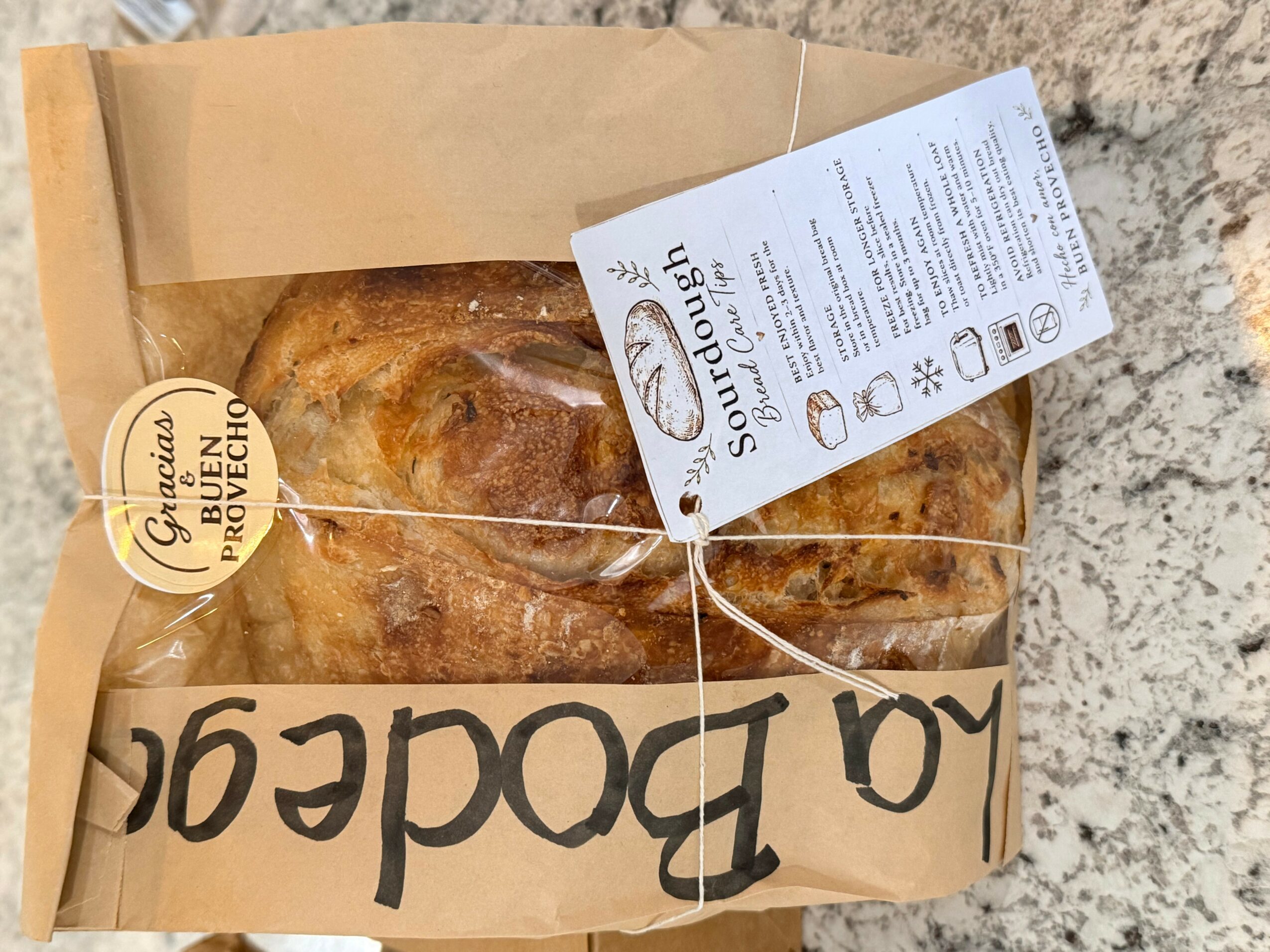 A loaf of bread in a paper bag with a handwritten label, a tag, and a sticker, on a textured surface.