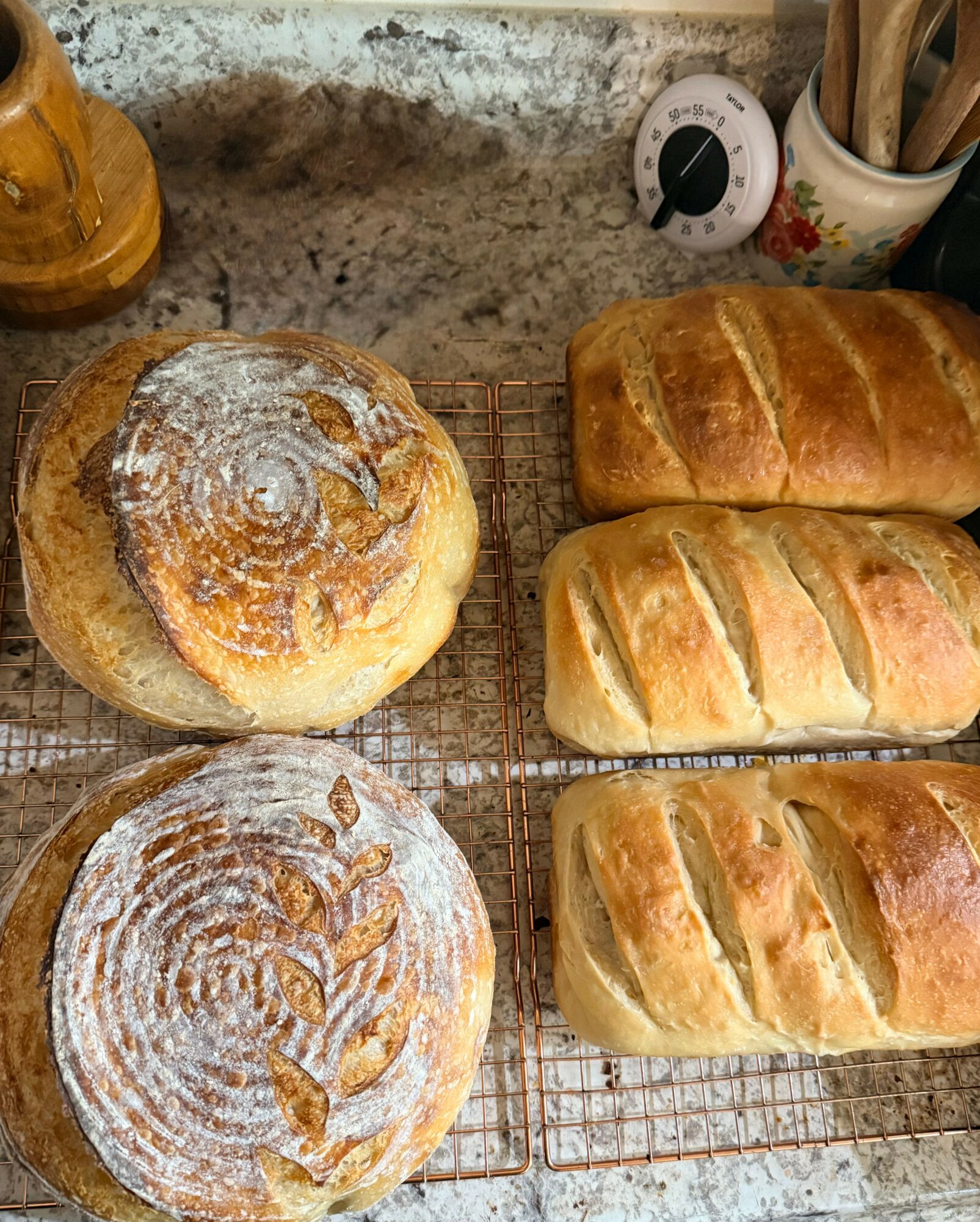 Three loaves of bread on a cooling rack, two with diagonal slashes, one round with a dusting of flour, on a kitchen counter.