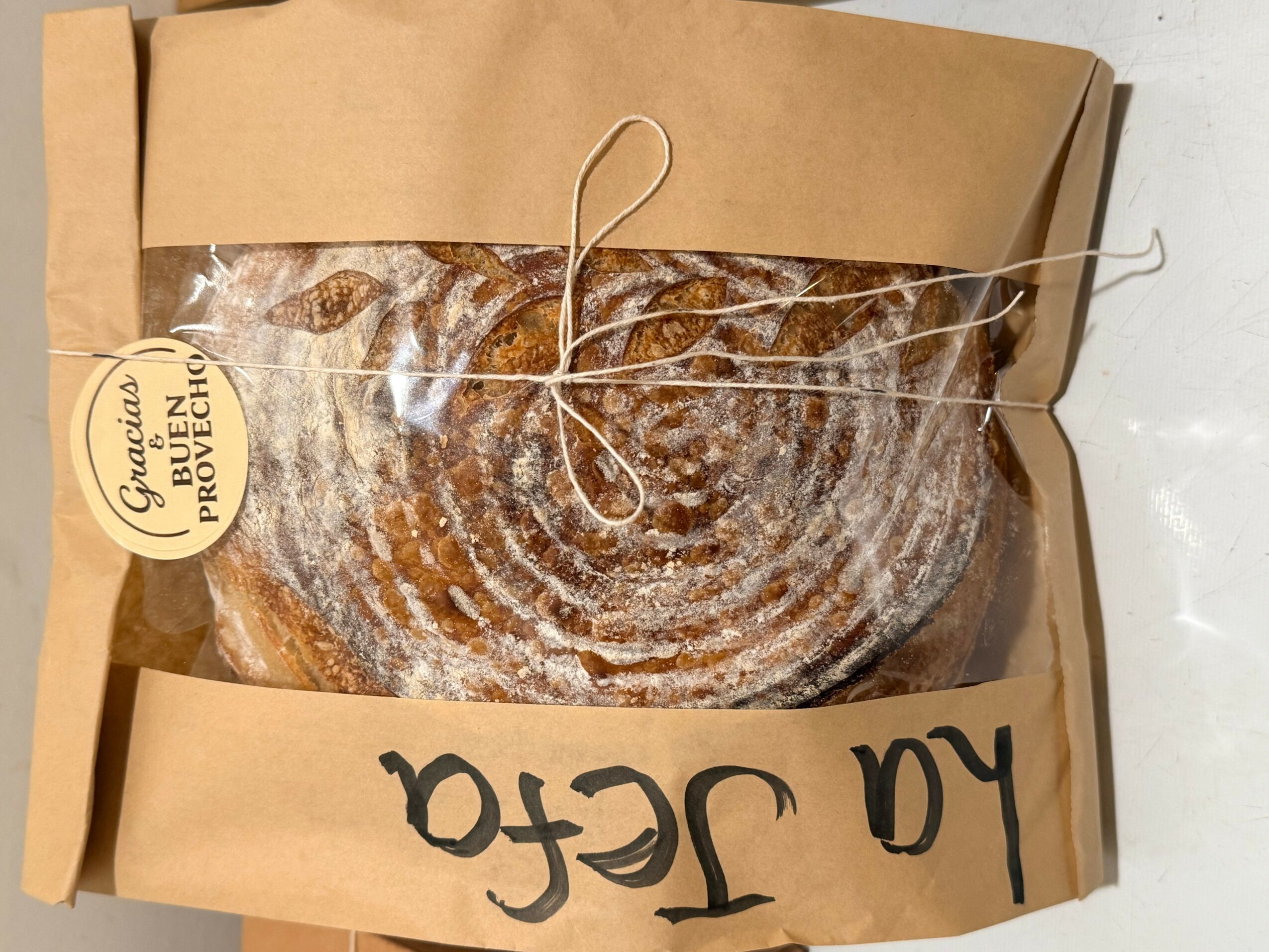 Round pastry with powdered sugar in a paper box, with a handwritten label and a sticker, viewed from above.