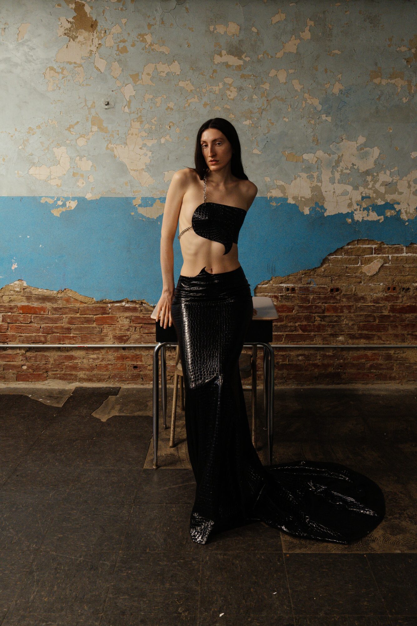 Woman in black strapless top and shiny black pants stands in front of a distressed wall with peeling paint, holding a bag.