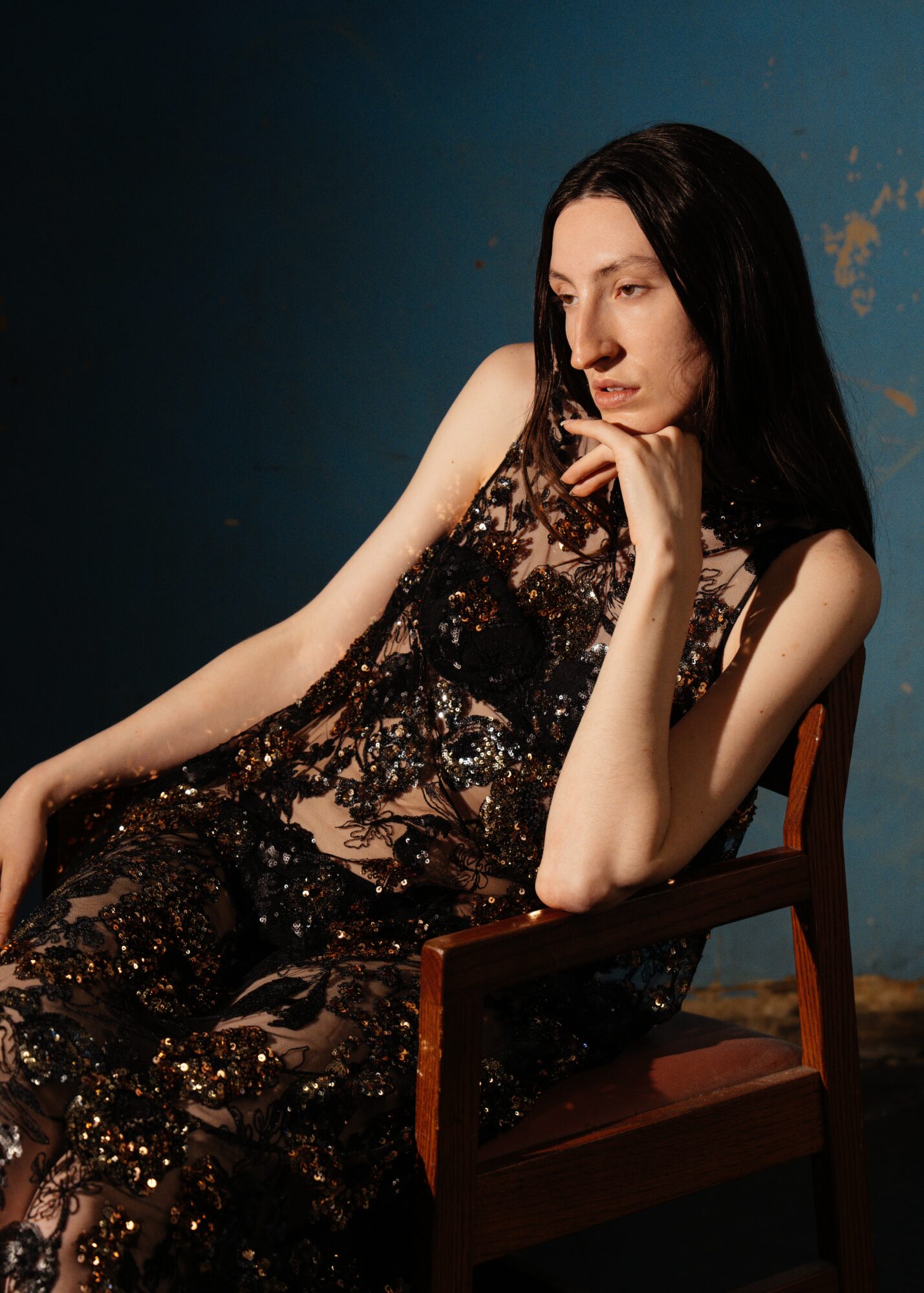 Woman with long dark hair sitting on a wooden chair, wearing a black sequined dress, against a blue wall.