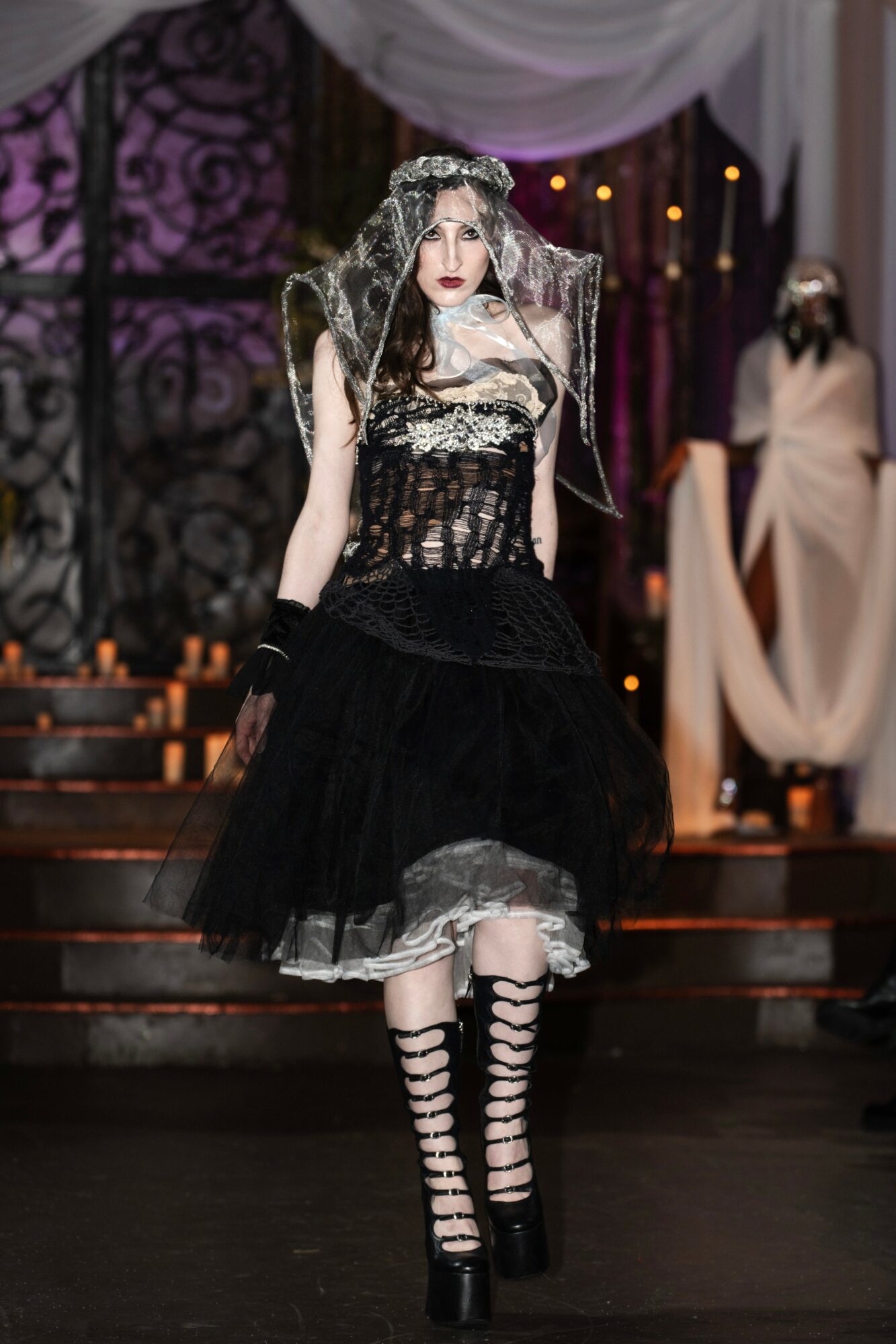 Person in gothic costume with veil, lace, and striped stockings on stage with draped curtains and another figure in background.