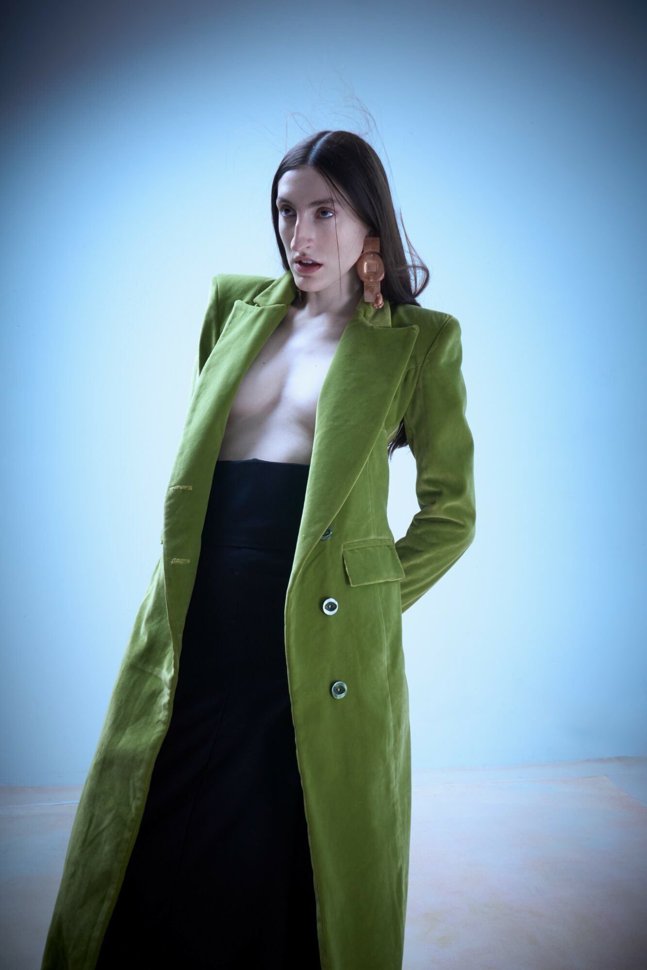 Woman with dark hair wearing a green coat over a black dress, standing against a light blue background.