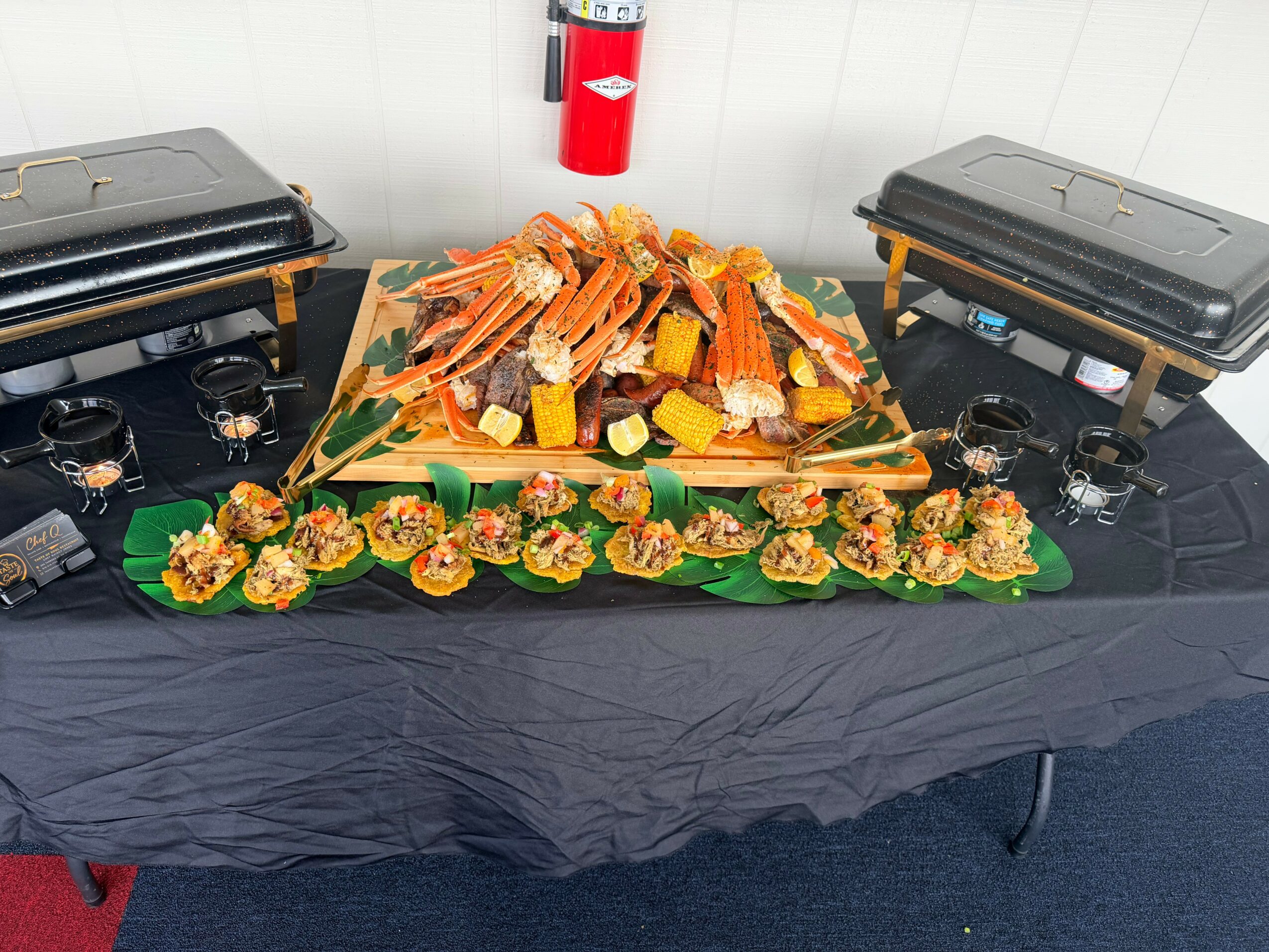 Food display with seafood, vegetables, and appetizers on a table with black tablecloth, two chafing dishes, and a fire extinguisher in the background.