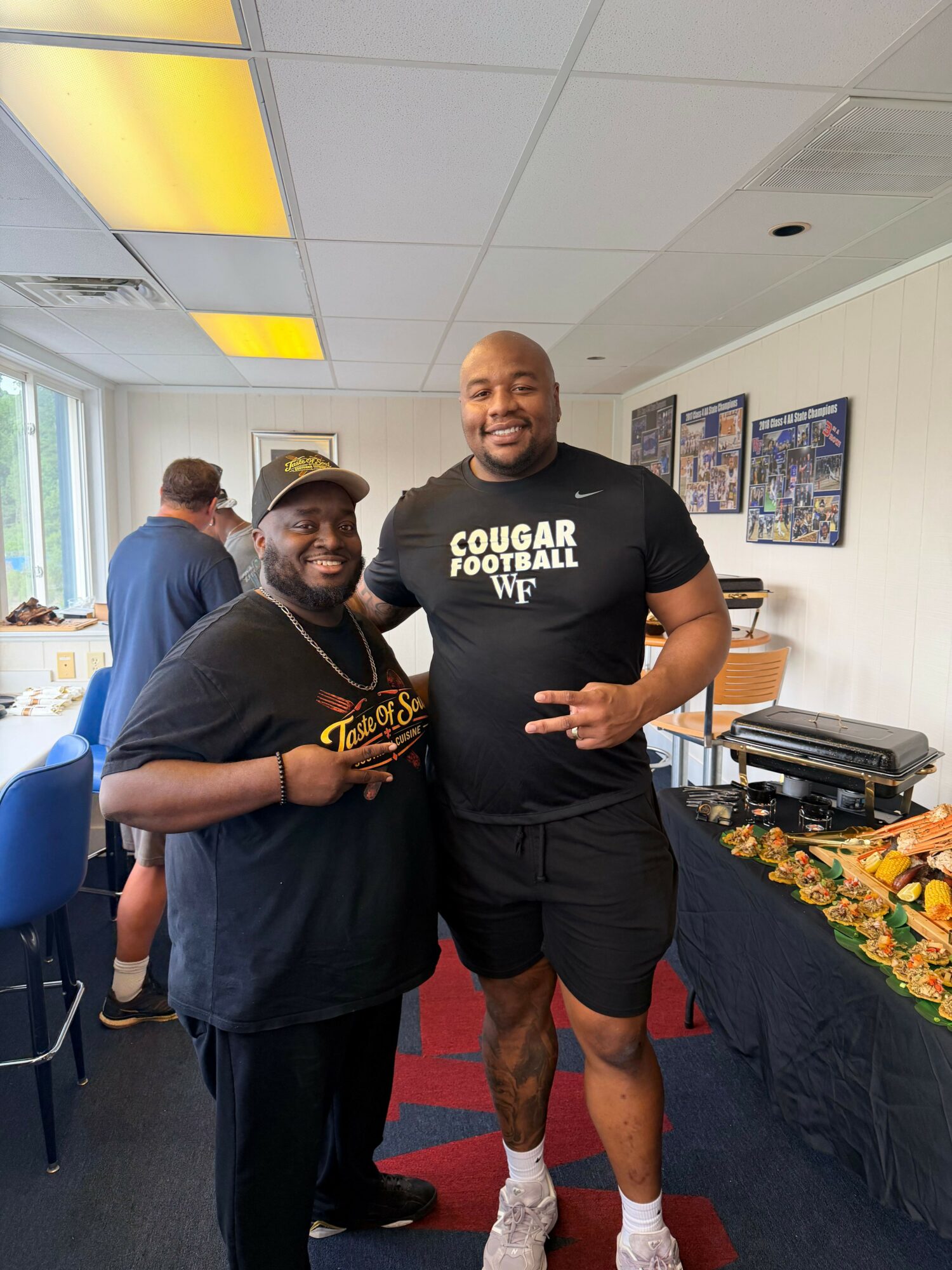 Two women standing together indoors, one wearing a black shirt and the other in a dark top, with a table of food behind them.