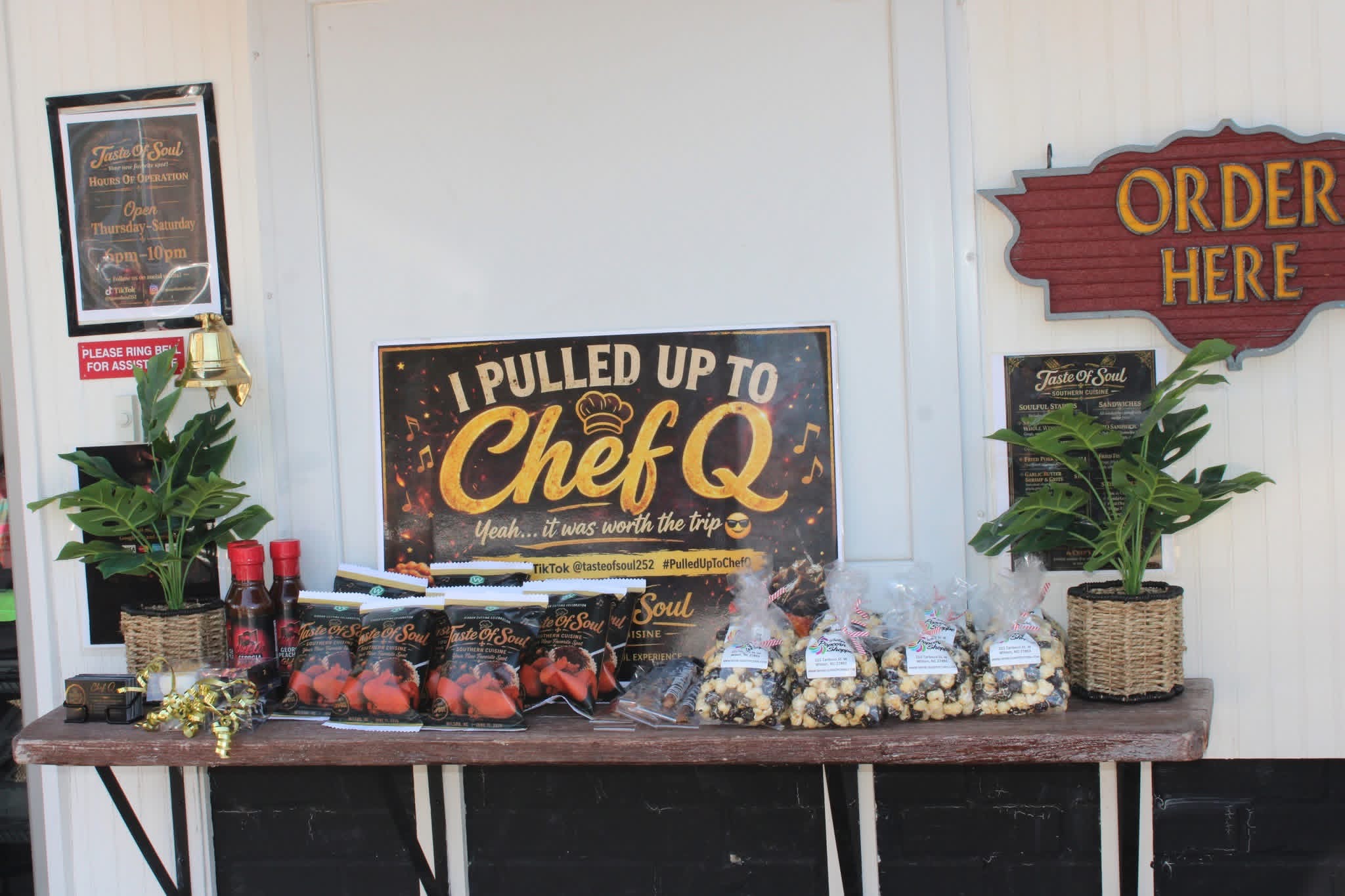 Table with food, drinks, and snacks, two potted plants, and signs in the background.