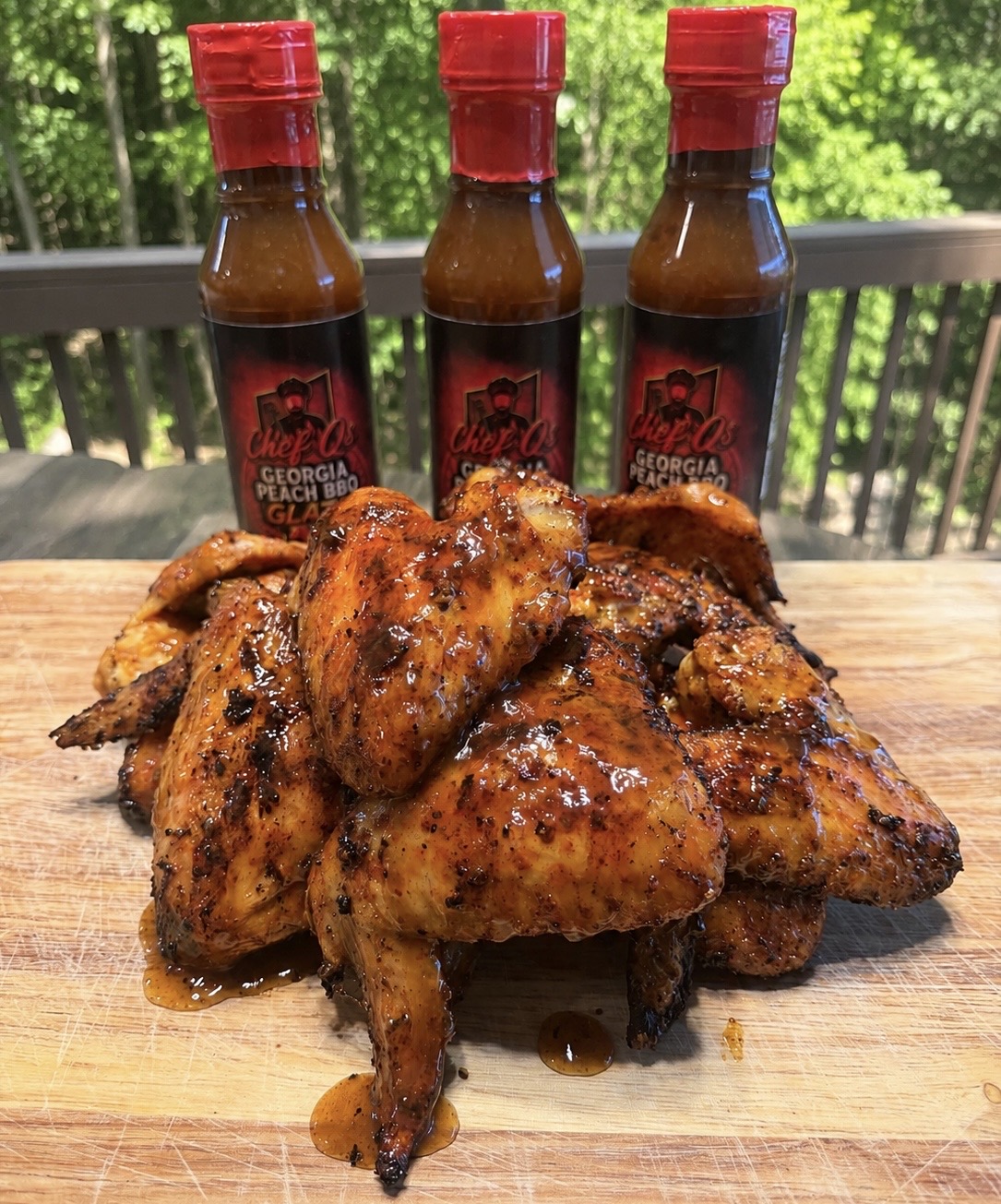 Barbecue chicken wings with three bottles of hot sauce on a wooden surface outdoors, with a green background.