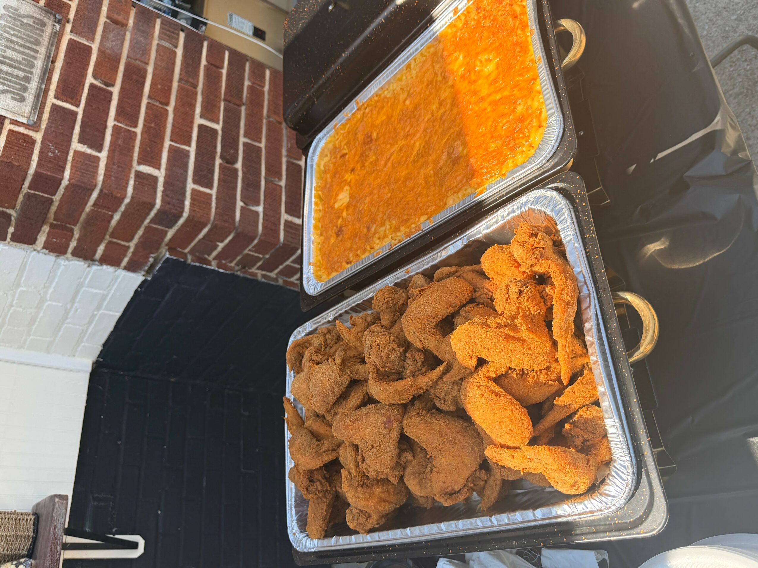 Two aluminum trays of fried chicken and a container of orange-colored sauce on a black surface.