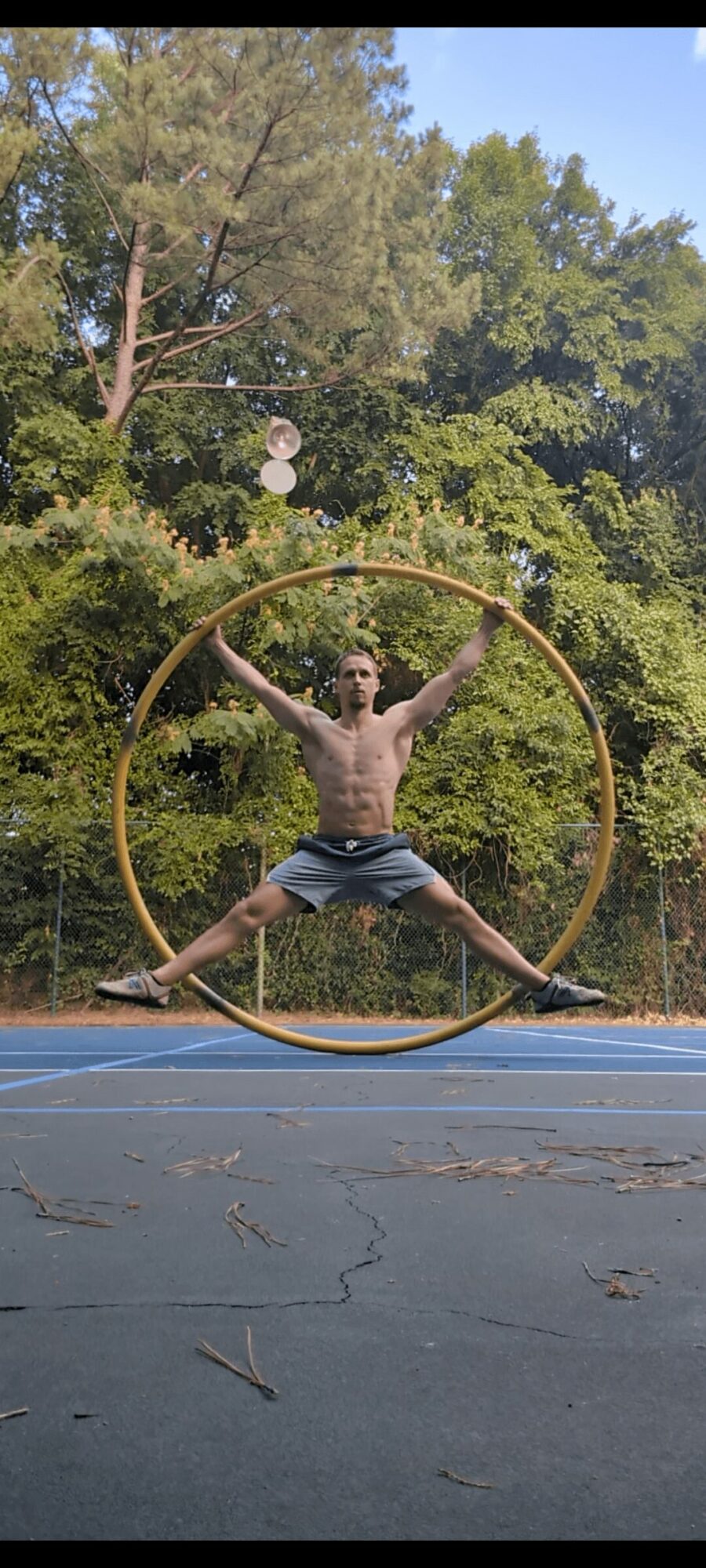 Person performing a split inside a large hula hoop outdoors, with trees in the background.