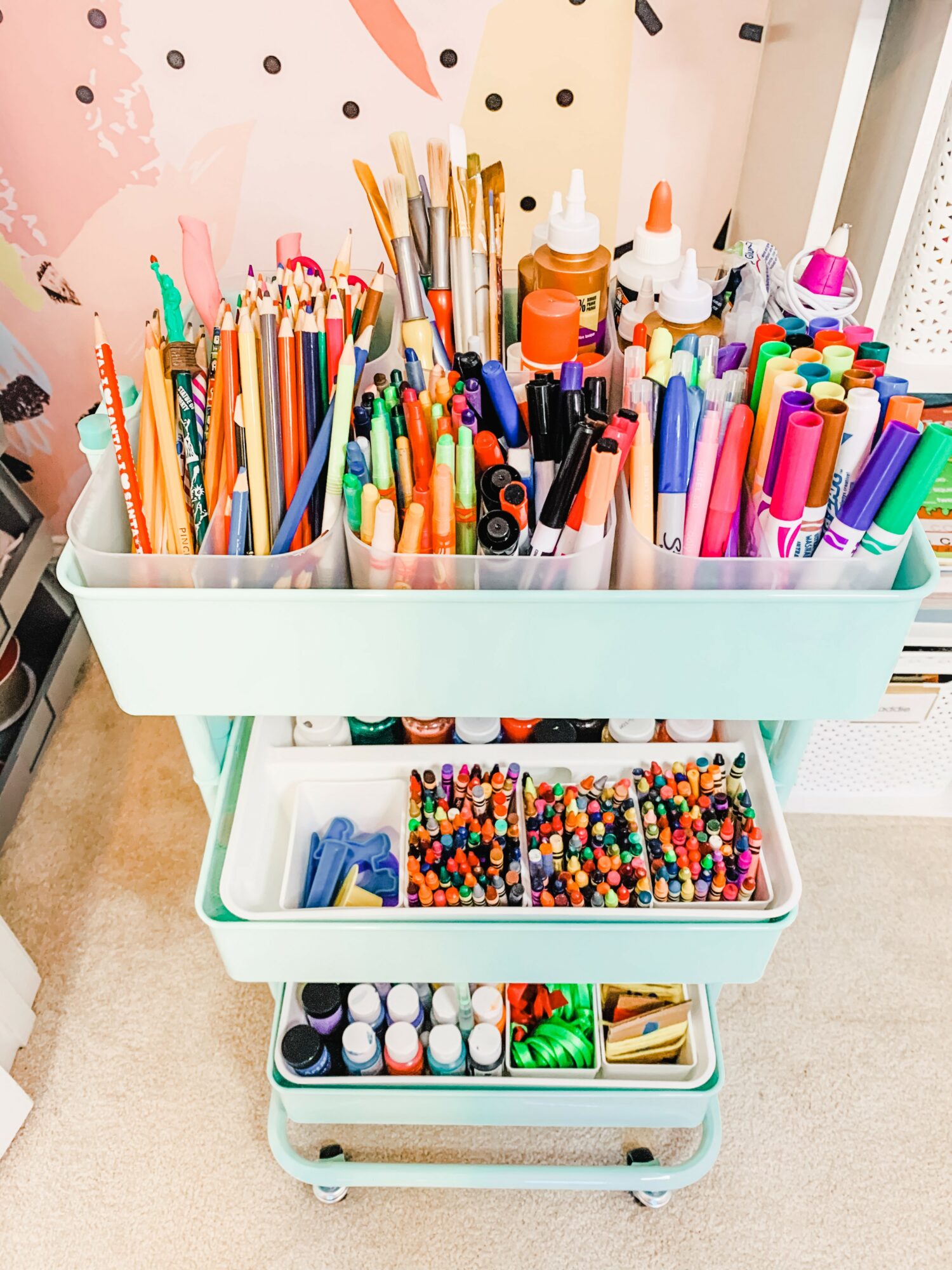 Cart filled with colorful markers, pens, and art supplies on a white trolley, with a pink wall in the background.