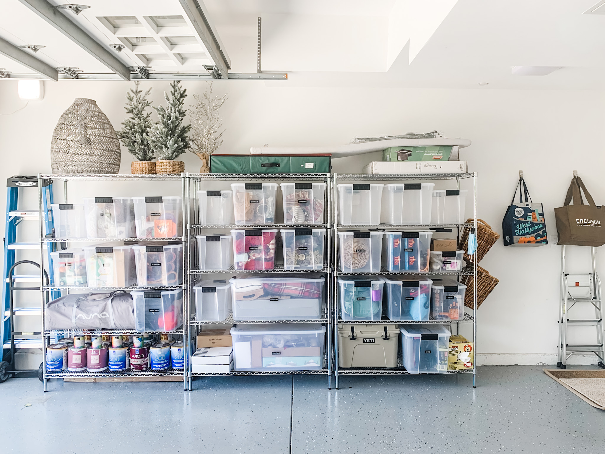 Shelving unit with plastic storage bins, boxes, and supplies in a bright room with white walls and a ladder.