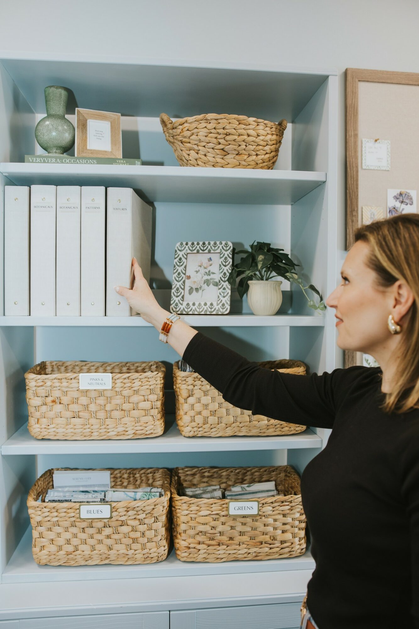 Woman reaching for a white binder on a light blue bookshelf with baskets and decorative items, side view, indoor setting.