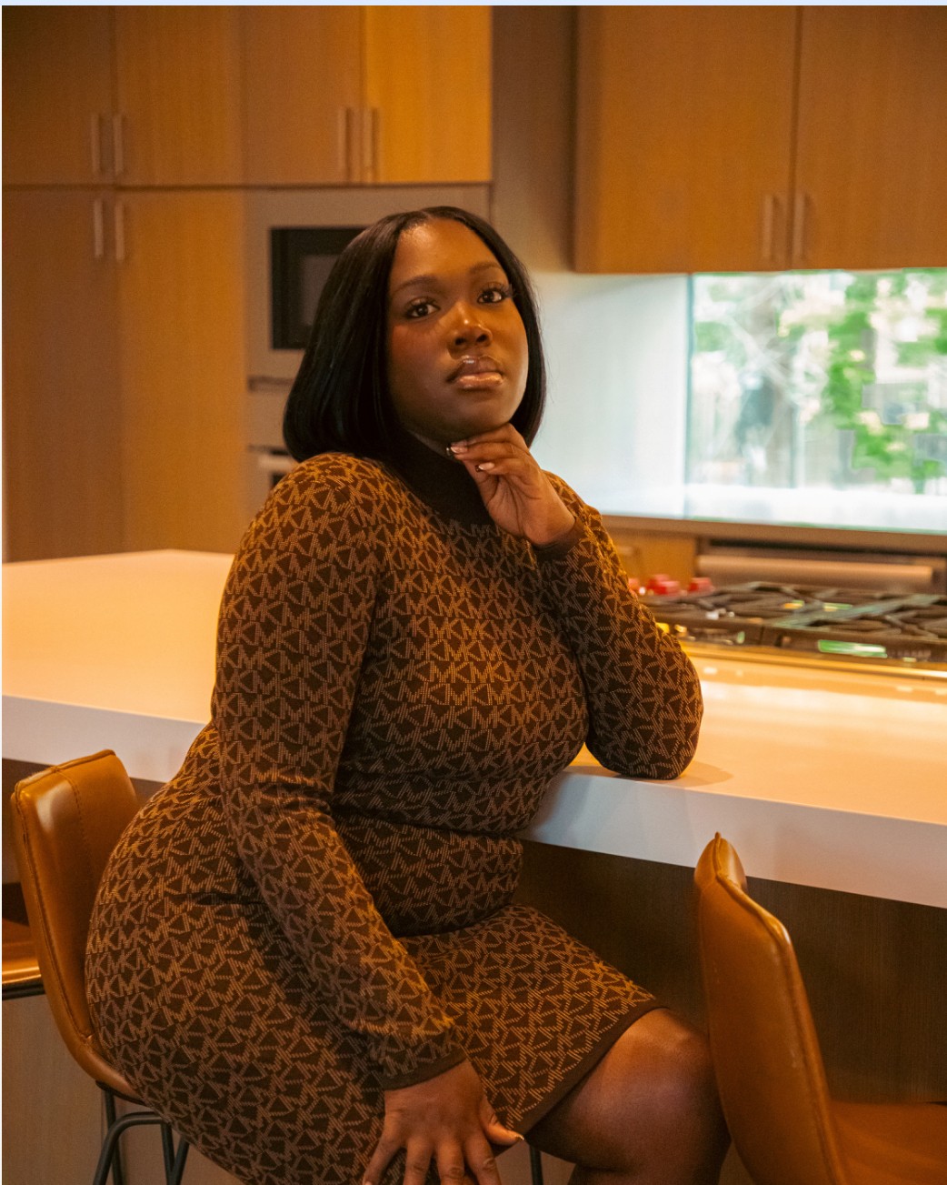 Woman with shoulder-length black hair sitting at a kitchen counter, wearing a patterned dress, with a window and wooden cabinets in background.