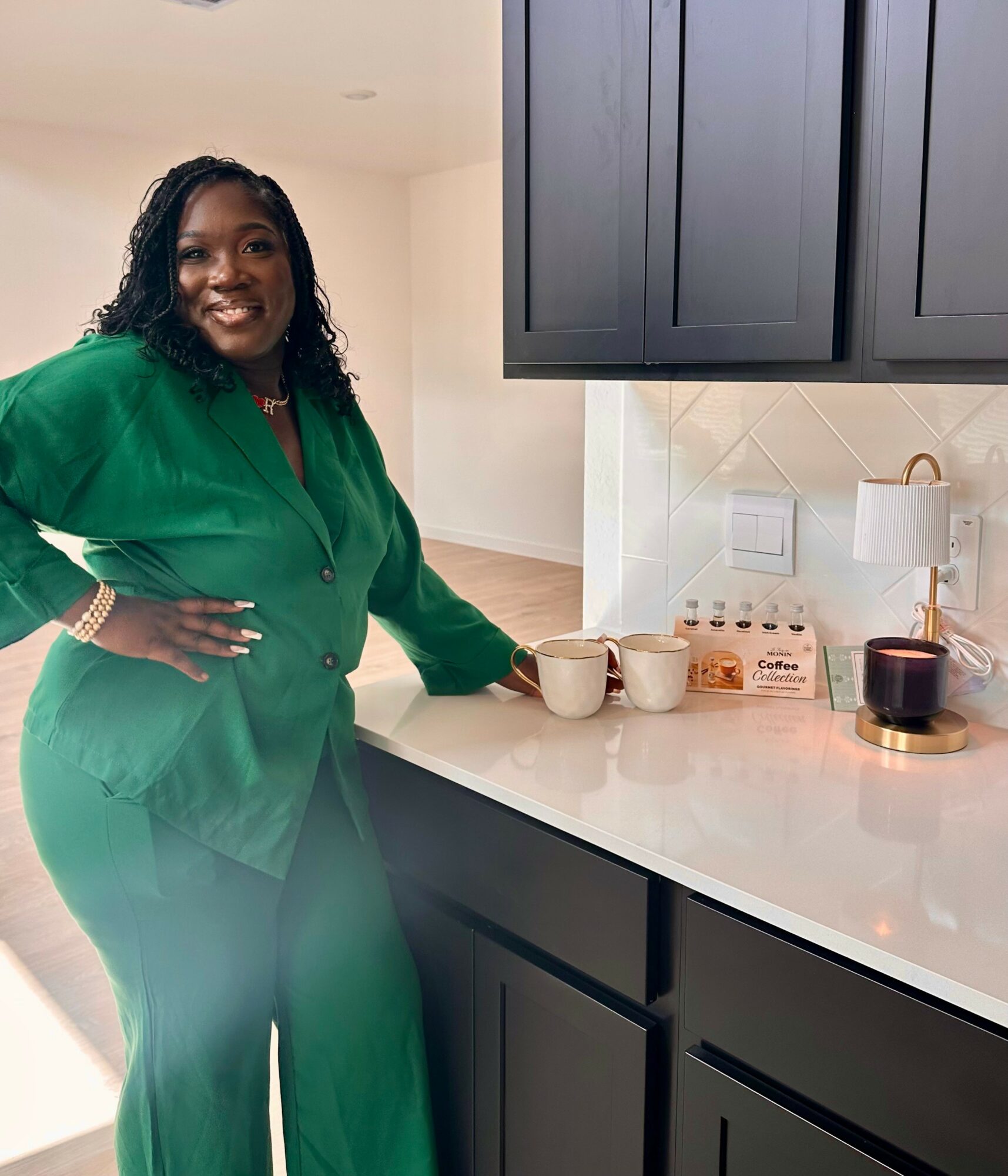 Woman in green outfit smiling, standing in a modern kitchen with dark cabinets and white countertop, holding mugs.