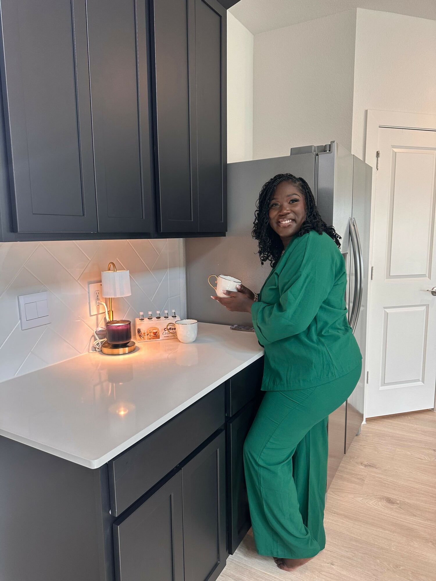 Woman in green scrubs leaning on a white table in a room with black and white cabinets, holding a cup.