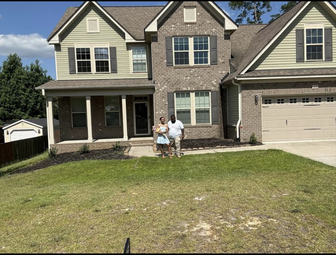 Two people standing in front of a two-story house with a garage, porch, and multiple windows, on a grassy lawn.