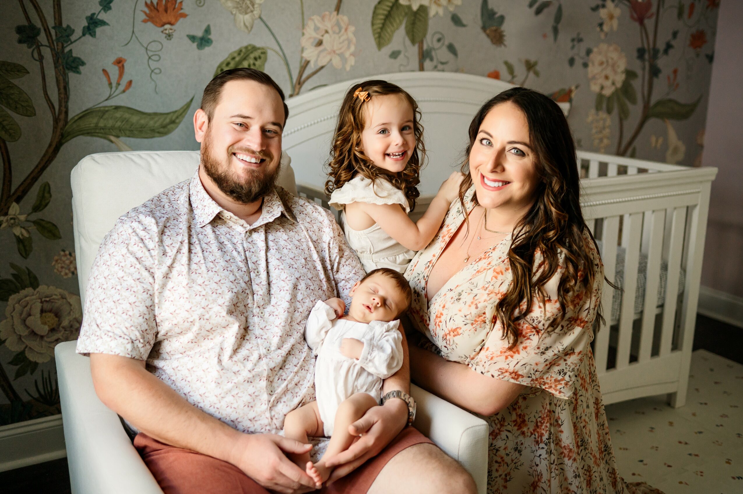Family of four with a newborn, sitting on a white chair, smiling, in a room with floral wallpaper.