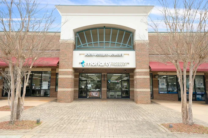 Building entrance with glass doors, brick facade, and trees on both sides, under a blue sky with clouds.