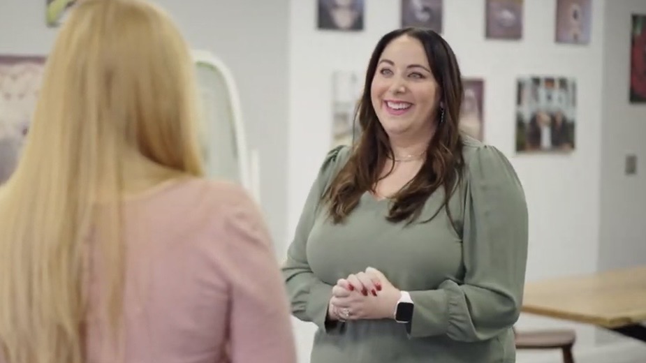 Two women talking indoors, one smiling, the other with back to camera, in a room with pictures on the wall.