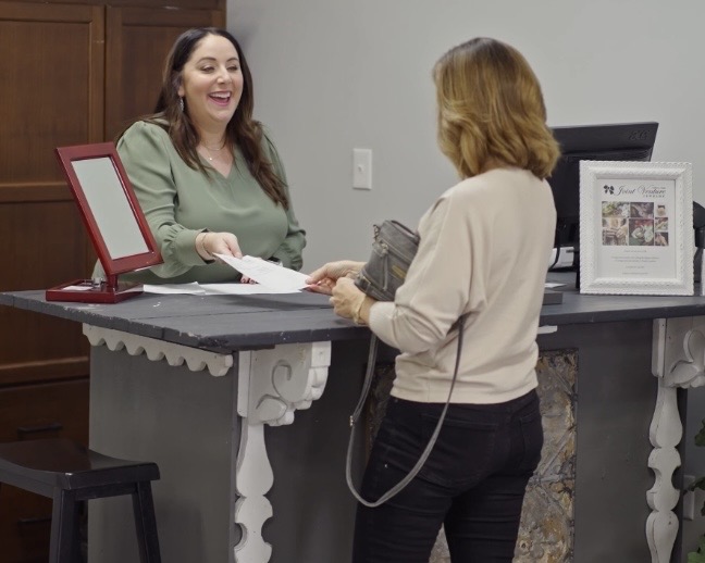 Two women at a reception desk exchanging documents, one smiling, in an indoor setting.