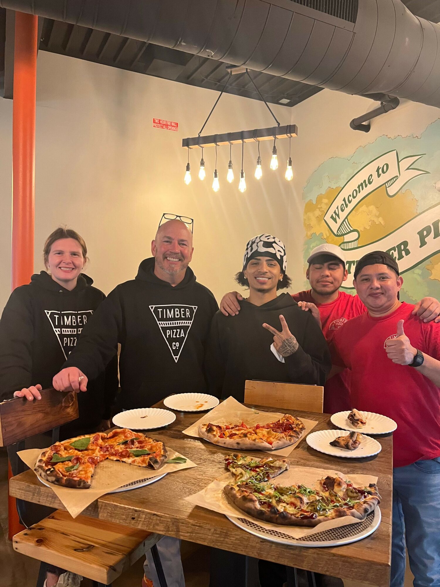 Four people standing behind a table with pizza, smiling at the camera in a restaurant.
