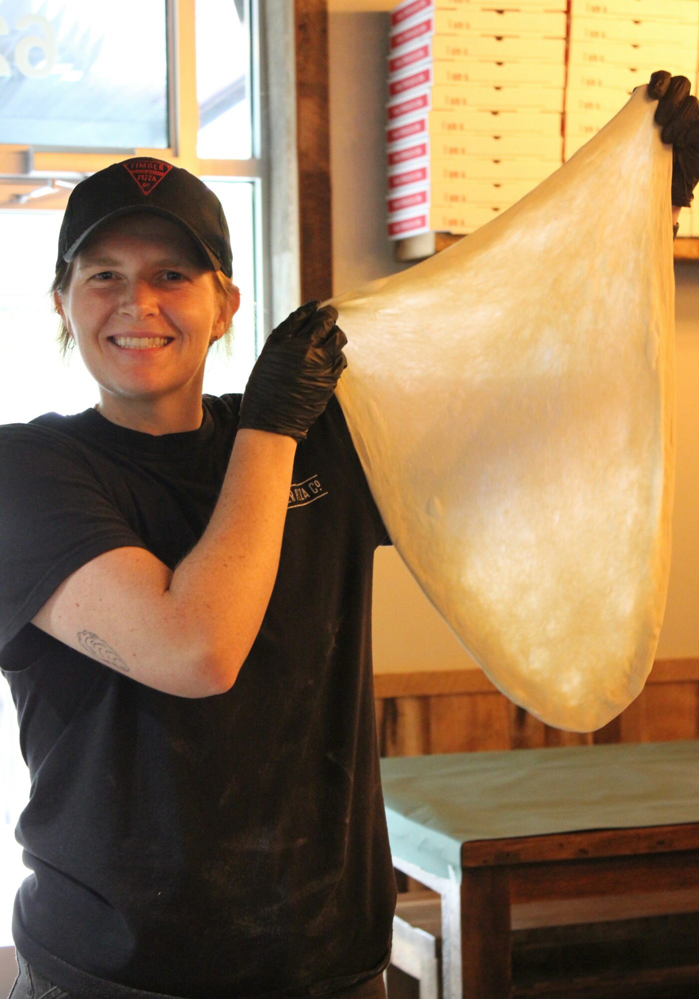 Person smiling, wearing black gloves and a cap, holding a large piece of dough in a bakery or kitchen setting.