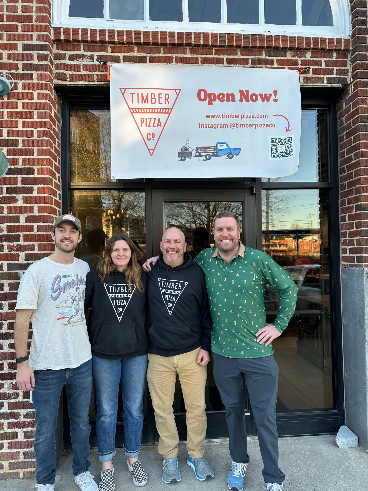 Four people stand outside a brick building with a glass door and a sign that reads 'Open Now!'.