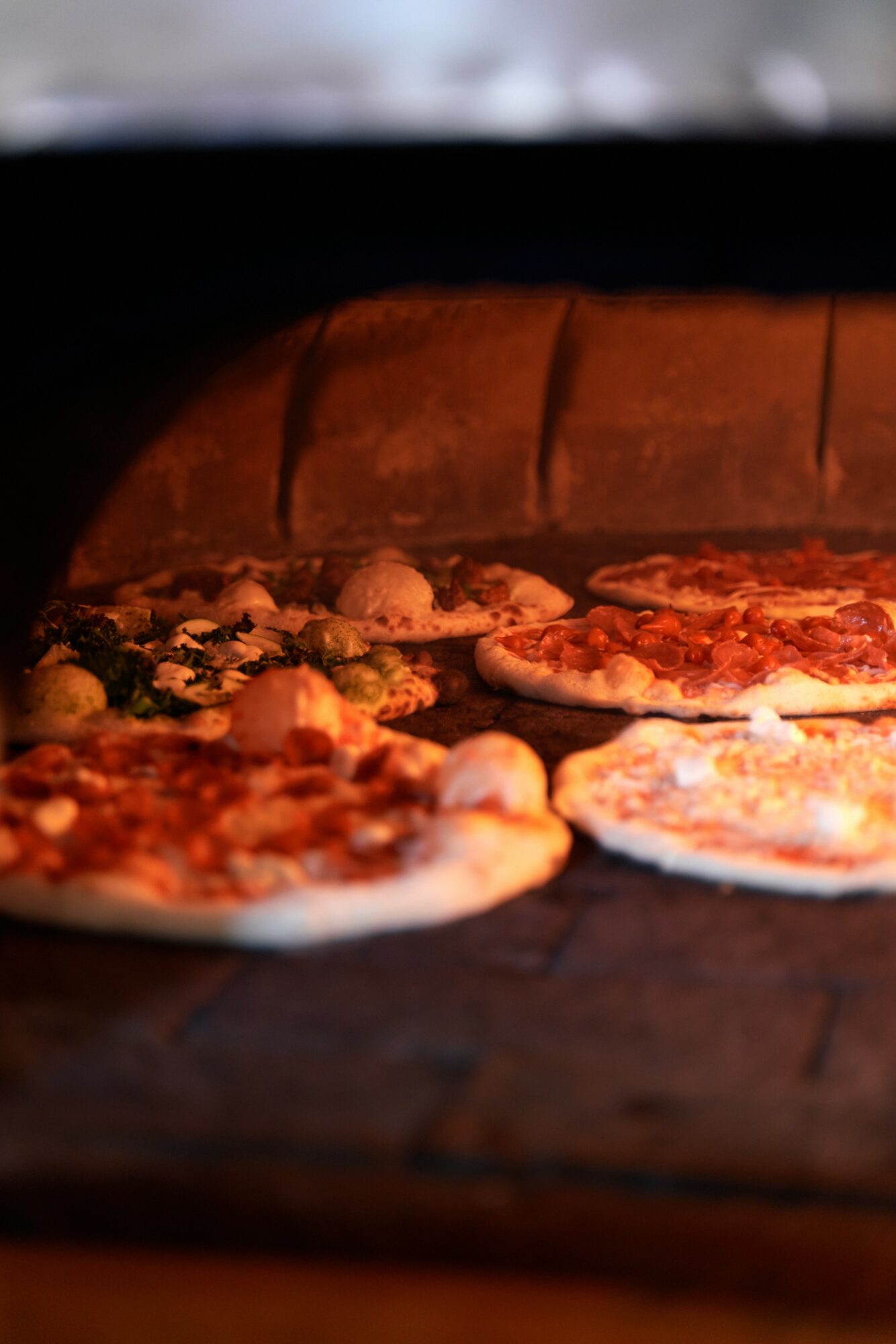Pizzas baking inside a wood-fired oven with a brick arch, visible toppings, and glowing heat.