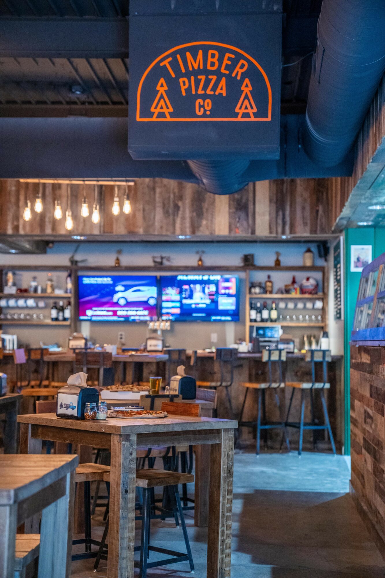 Interior of a restaurant with wooden tables, chairs, and a bar area with digital menu screens, shelves with bottles, and a hanging sign.