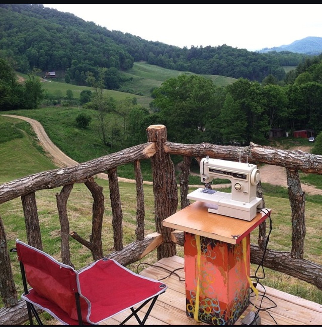 A sewing machine on a small table on a wooden balcony with a scenic green landscape and hills in the background.