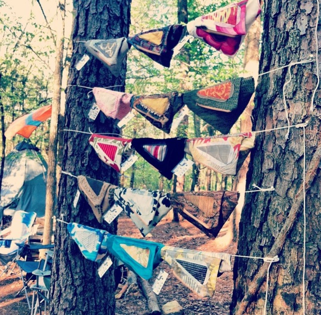 Colorful fabric bunting hanging between two trees in a forest setting, with trees and greenery in the background.